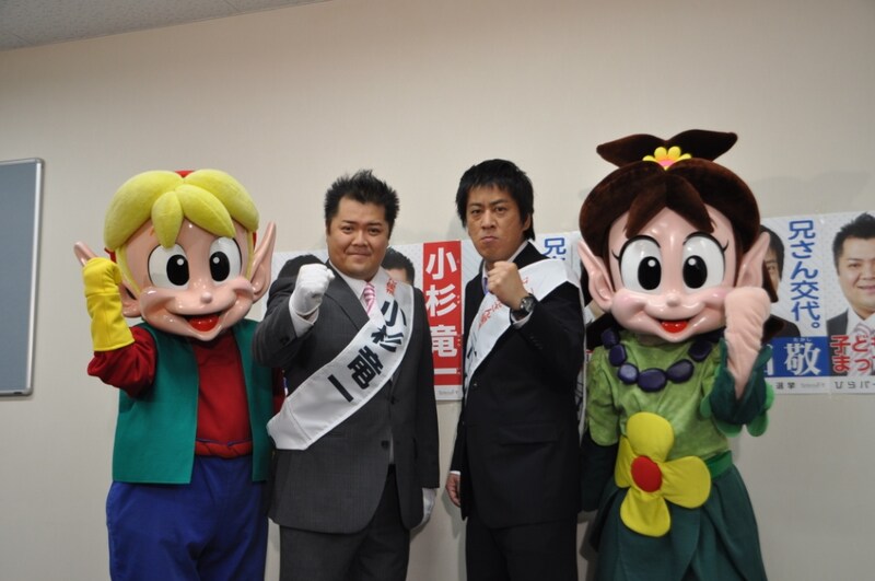 2代目ひらパー兄さん選挙で戦う現職の小杉竜一と新人の吉田敬。両隣は2人を応援するひらパーキャラクター・ピピンハーキン（左）とポピーハーケン（右）。