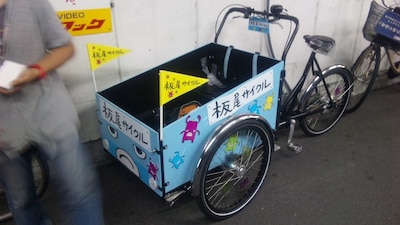 板尾が乗った自転車。前の箱部分に人が乗り、板尾が後ろから話かける形になる。