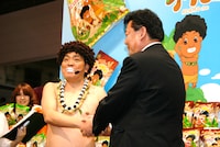 「アダモちゃん ひねりスナック」のパッケージの可愛さと美味しさに太鼓判。この菓子をプロデュースした会社の上役と握手を交わすアダモちゃんこと島崎俊郎。