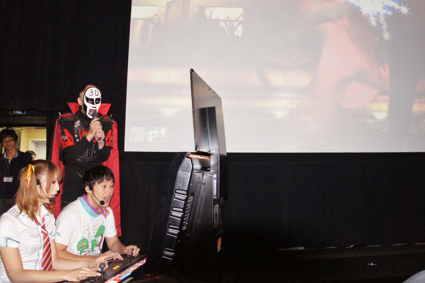 今秋稼働予定のアーケードゲーム「鉄拳タッグトーナメント2」で対戦する佐藤かよと佐久間一行（左手前から）。劇場のスクリーンにもゲーム画面が映し出され、観客を盛り上げた。