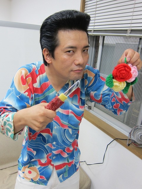 趣味の編み物展示をハブ展にて行うえんにち・アイパー滝沢。手には毛糸のカバーが付いた匕首と、バラがモチーフのアクリルたわし。