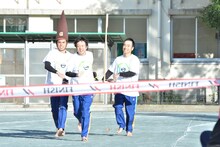 裸足で校庭を走る、小島よしおとかもめんたる（左から）。写真は靴を運ぶレース「はだしラン」のデモンストレーションの様子。