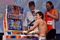 秋山（右）と馬場（左）にそそのかされ、データカードダスの前でキーボーディストを演じる山本（手前）。