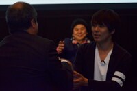 シネマサンシャインを展開する佐々木興業株式会社・佐々木社長（左）に握手を求めるウーマンラッシュアワー村本（右）。