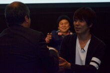 シネマサンシャインを展開する佐々木興業株式会社・佐々木社長（左）に握手を求めるウーマンラッシュアワー村本（右）。