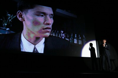 映画「ベイブルース～25歳と364日～」の舞台挨拶で、河本栄得の映像を流しながらネタを披露する高山トモヒロ監督（右）。