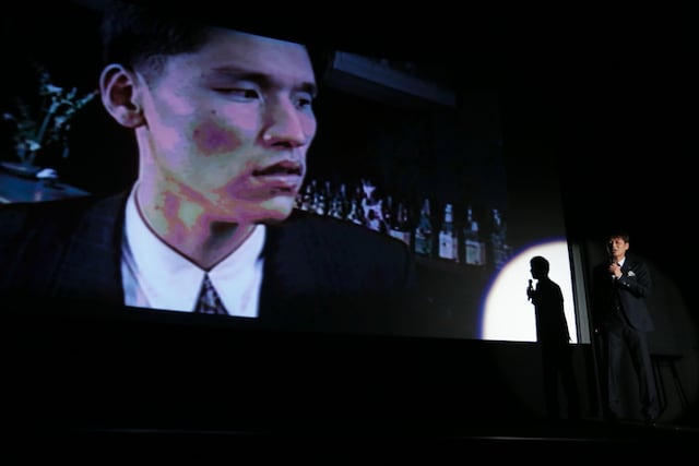 映画「ベイブルース～25歳と364日～」の舞台挨拶で、河本栄得の映像を流しながらネタを披露する高山トモヒロ監督（右）。