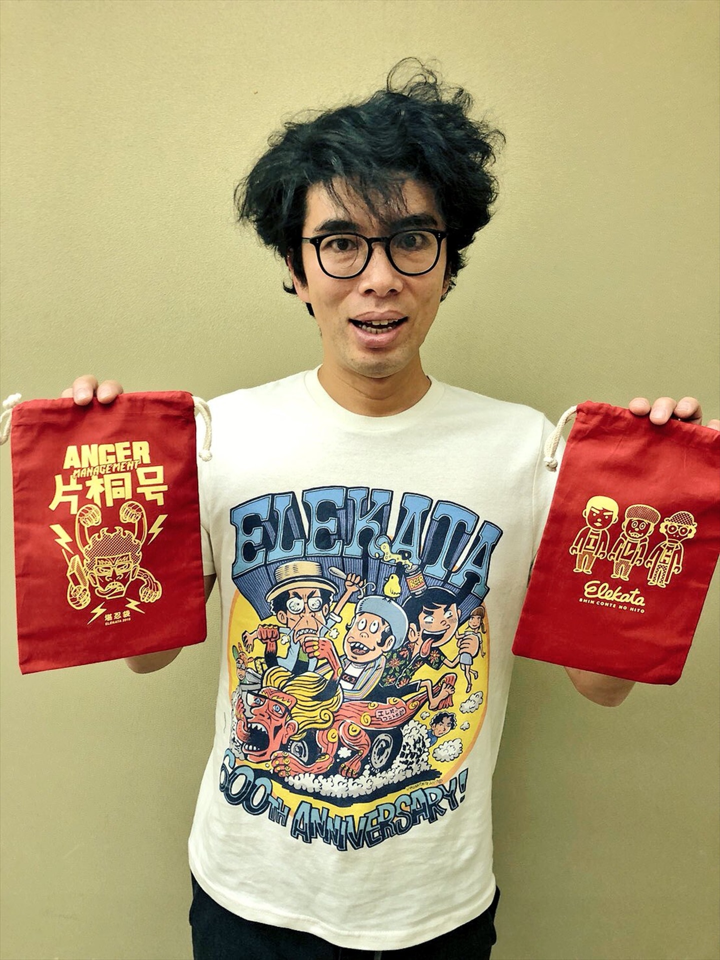 グッズのTシャツを着用し、巾着袋を持つ片桐仁。