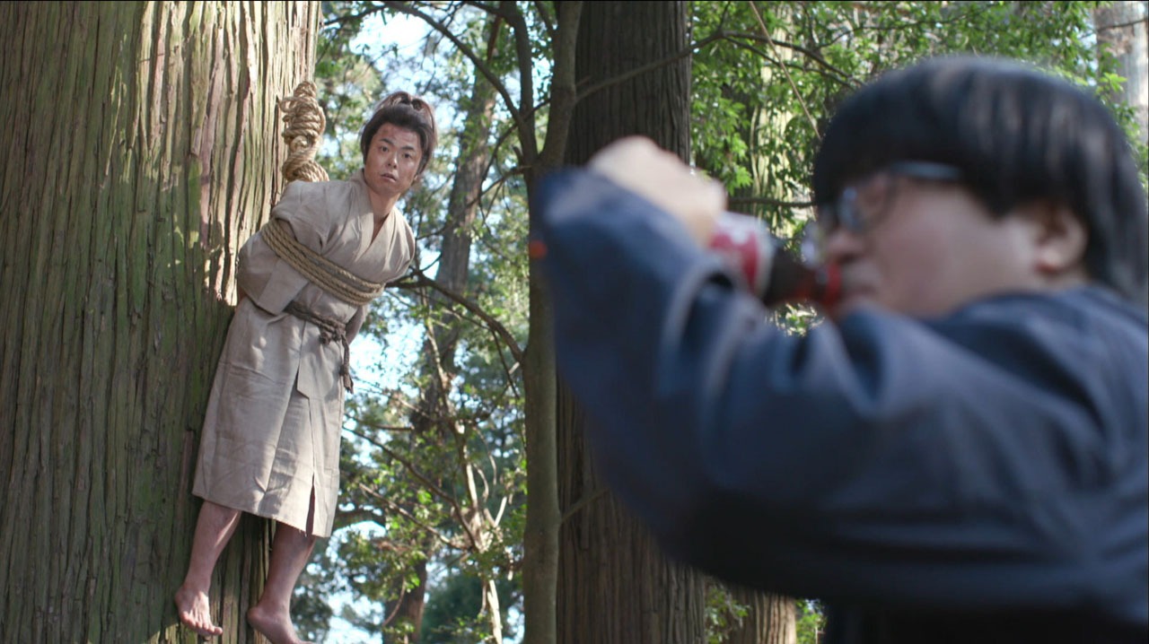 宮本武蔵役の上川隆也に扮し、大木に吊るされるライス田所（左）と、コーラを飲むAD役のライス関町（右）。(c)テレビ東京