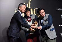 西川昌宏編集長からトロフィーを受け取るミキ昴生（右）と2人の間からひょっこり顔を出す亜生。