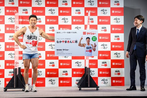 “やきにくのタレ営業部長”の巨大名刺を左腕の筋肉だけで持つなかやまきんに君（右）。
