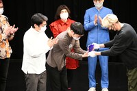 ゲスト審査員のPOISON GIRL BAND吉田からトロフィーを受け取る男性ブランコ。