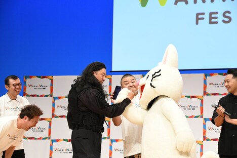 チンギスと公安・野崎のように握手を交わすレイザーラモンRGとノンタン、うしろはスマホの翻訳アプリを見せるドラム役の川畑泰史。