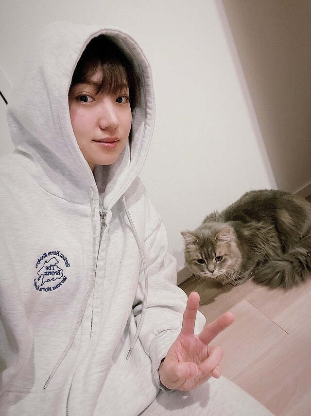 活発な印象の、ロゴ入りのパーカ姿でピース。後ろは太田のピースに釘付けの愛猫。