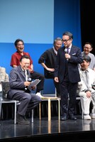 自分だけ違う種類のいすに座ることになり、納得がいかない様子の三谷幸喜（前列右）。