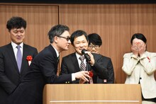 森下創（中央）があいさつしている最中、マイクの高さ調整を手伝う安井順平（左から2番目）。