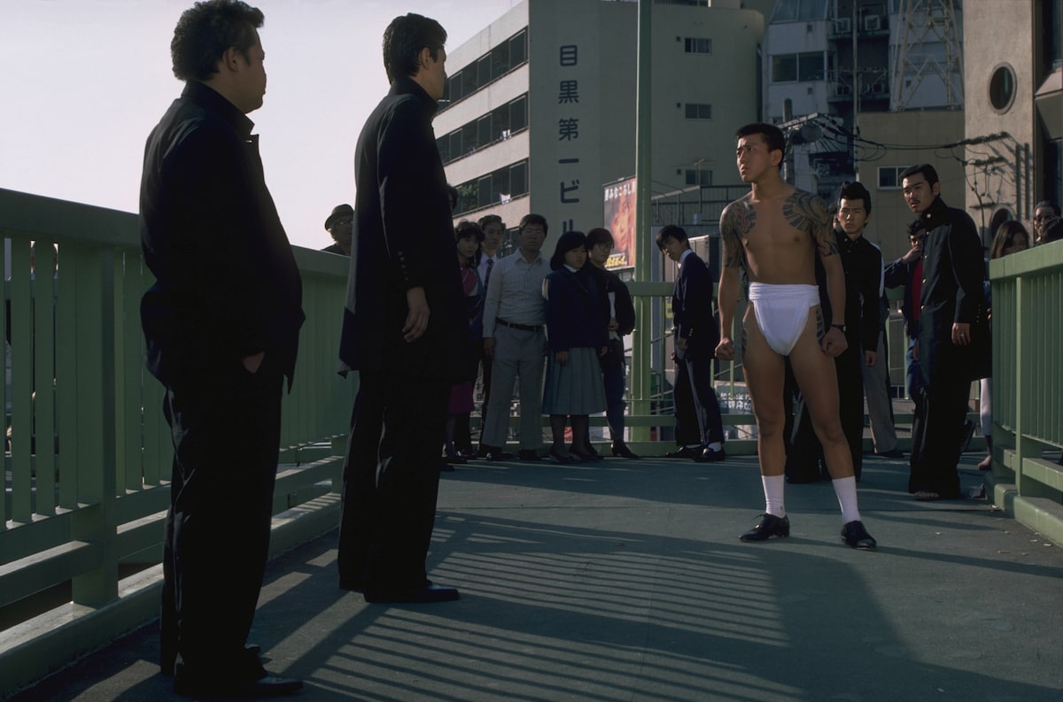 映画「ビー・バップ・ハイスクール」より。ヒロシとトオルの押しかけ舎弟であるノブオは、2人からのテストとして北高の前川新吾に突っかかる。歩道橋の上で追い詰められたノブオが脱いだ学ランは、下を通っていた車の視界をふさぎ、事故が起きる ©︎1985 東映 セントラル・アーツ